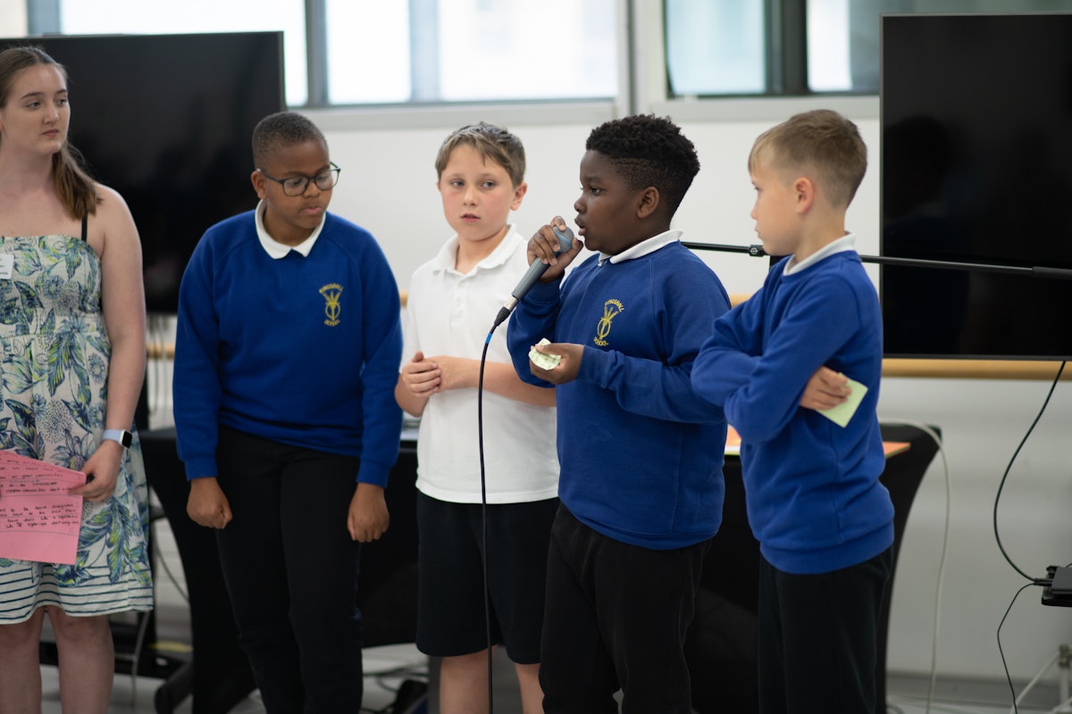 A group of primary school students in blue school uniforms stand together, with one student speaking into a microphone while presenting their ideas to the room